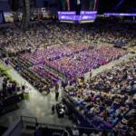 Wide view of a college graduation ceremony in an arena; hundreds of graduates in purple gowns sit in rows with a large audience surrounding them, stage on left.