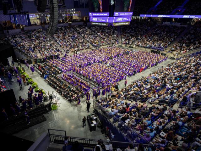 Wide view of a college graduation ceremony in an arena; hundreds of graduates in purple gowns sit in rows with a large audience surrounding them, stage on left.