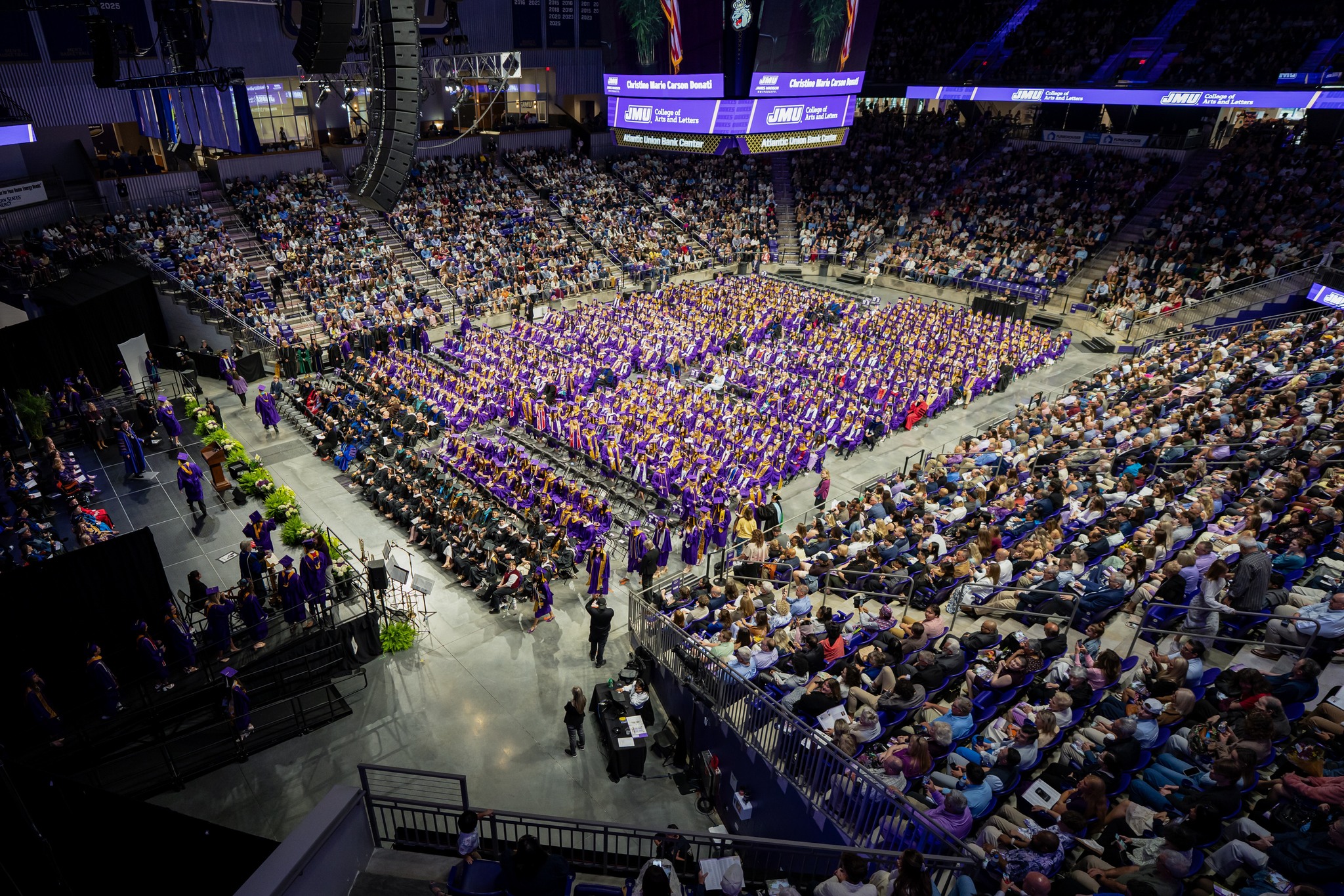 Wide view of a college graduation ceremony in an arena; hundreds of graduates in purple gowns sit in rows with a large audience surrounding them, stage on left.