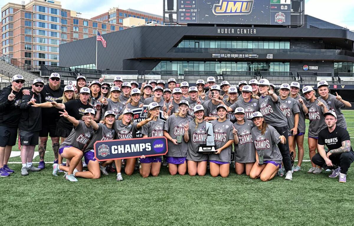 Women’s lacrosse team posing on the field with a ‘CHAMPS’ banner and trophy after winning a championship at a stadium.