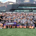 Women’s lacrosse team posing on the field with a ‘CHAMPS’ banner and trophy after winning a championship at a stadium.