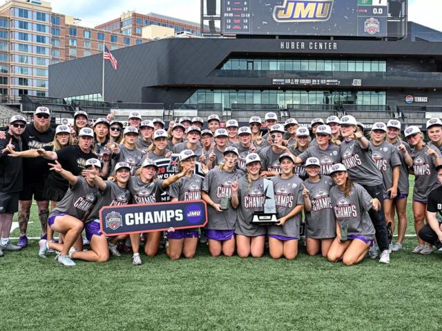 Women’s lacrosse team posing on the field with a ‘CHAMPS’ banner and trophy after winning a championship at a stadium.