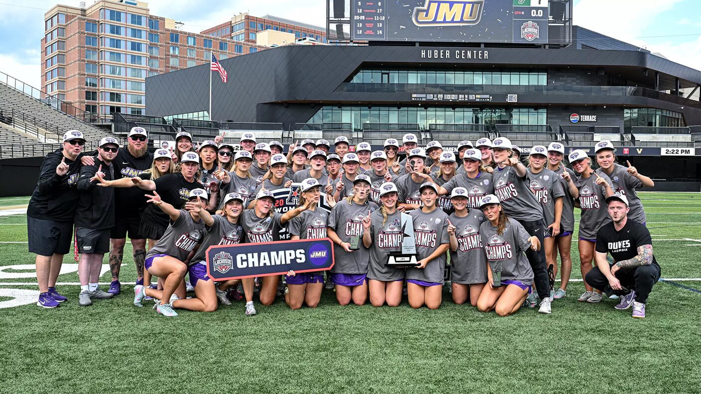 Women’s lacrosse team posing on the field with a ‘CHAMPS’ banner and trophy after winning a championship at a stadium.