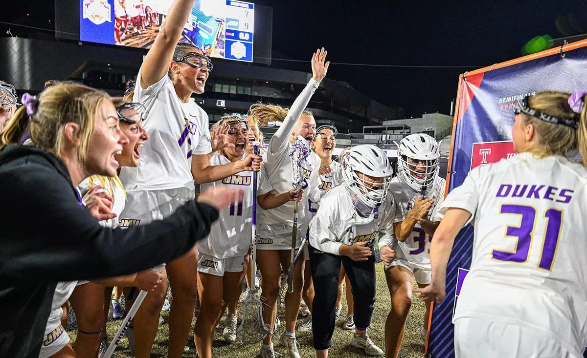 Dukes lacrosse team celebrates a victory on the field at night, players in white uniforms cheering and high-fiving.