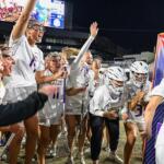 Dukes lacrosse team celebrates a victory on the field at night, players in white uniforms cheering and high-fiving.