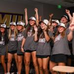 Group of young women in gray Champs shirts celebrate together in a locker-room, arms raised and smiling after a win.