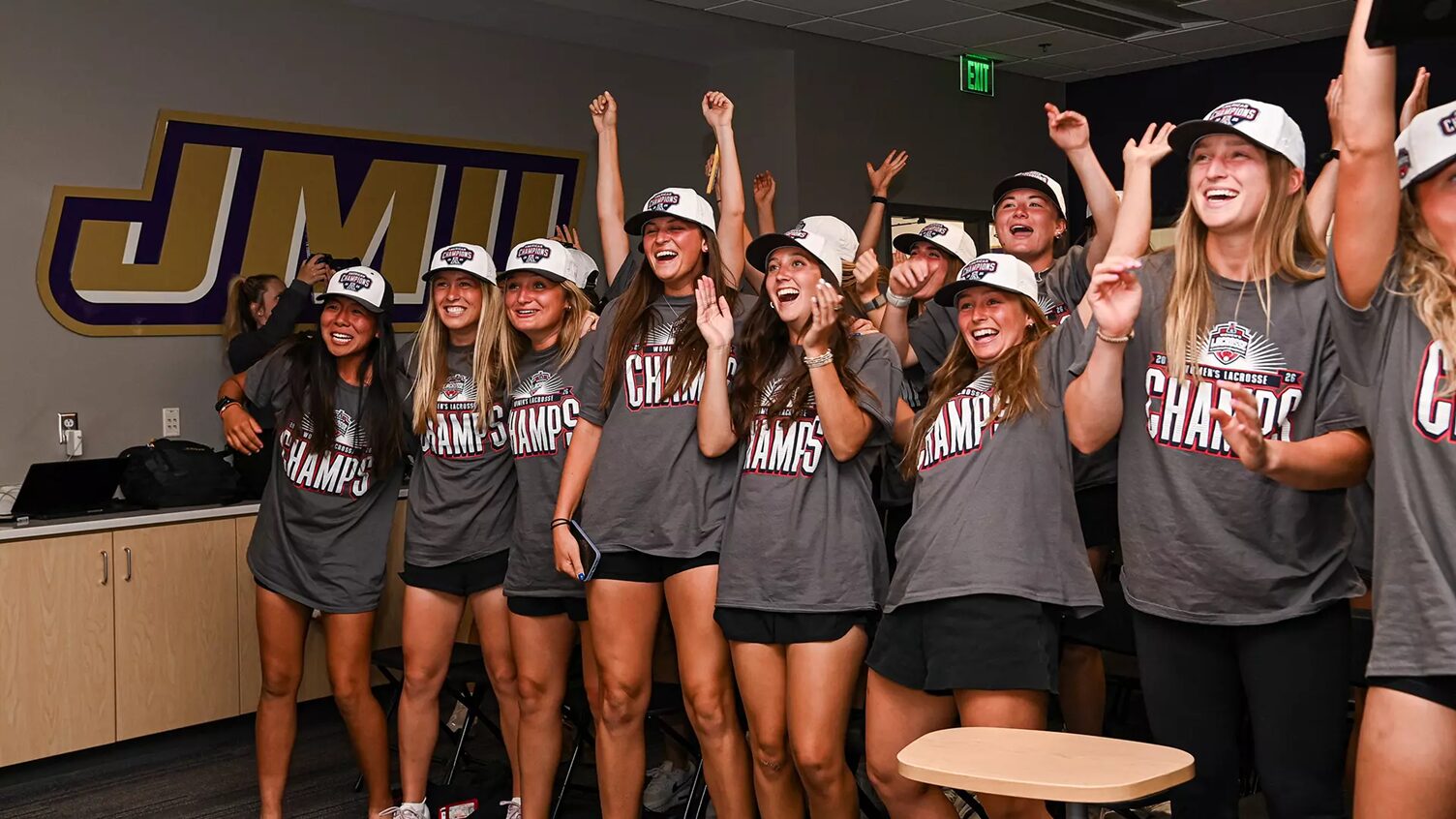 Group of young women in gray Champs shirts celebrate together in a locker-room, arms raised and smiling after a win.