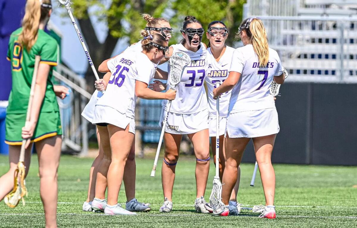 Girls' lacrosse team in white uniforms huddle with sticks on a sunny green field; jerseys show JMU and numbers.