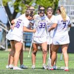 Girls' lacrosse team in white uniforms huddle with sticks on a sunny green field; jerseys show JMU and numbers.
