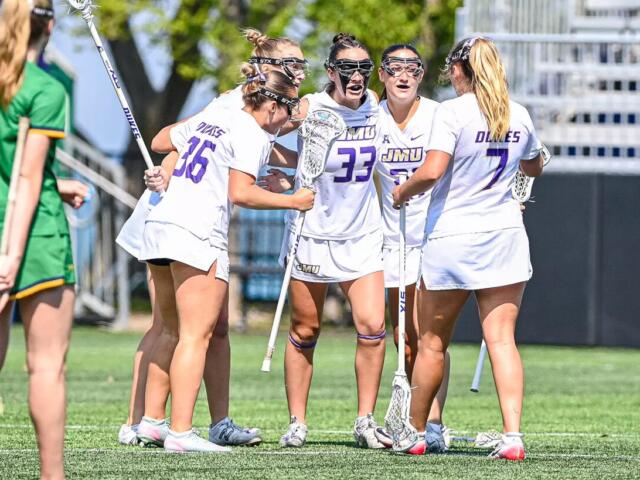 Girls' lacrosse team in white uniforms huddle with sticks on a sunny green field; jerseys show JMU and numbers.
