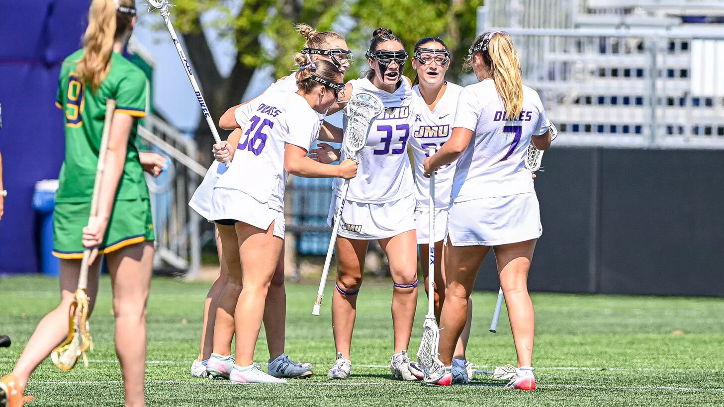 Girls' lacrosse team in white uniforms huddle with sticks on a sunny green field; jerseys show JMU and numbers.