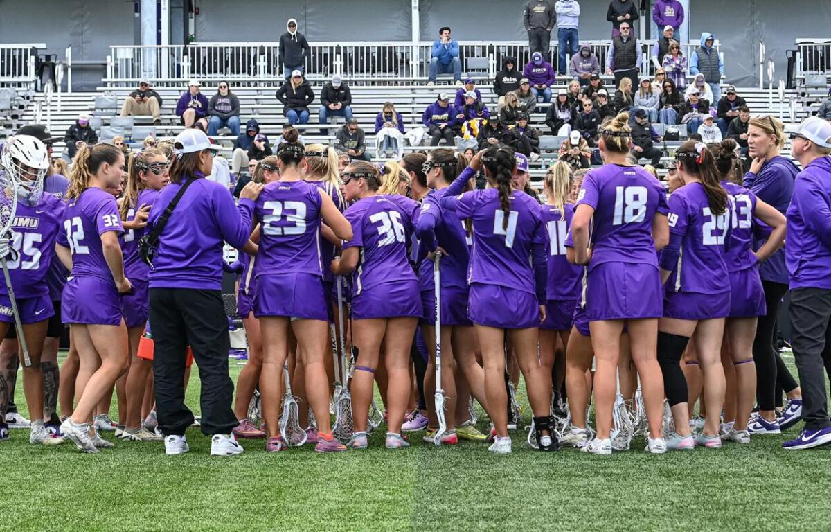 Lacrosse team in purple uniforms huddles on the field with sticks, players leaning in for a pep talk as spectators watch nearby.