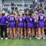 Lacrosse team in purple uniforms huddles on the field with sticks, players leaning in for a pep talk as spectators watch nearby.