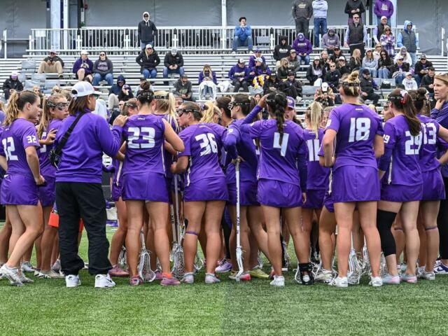 Lacrosse team in purple uniforms huddles on the field with sticks, players leaning in for a pep talk as spectators watch nearby.
