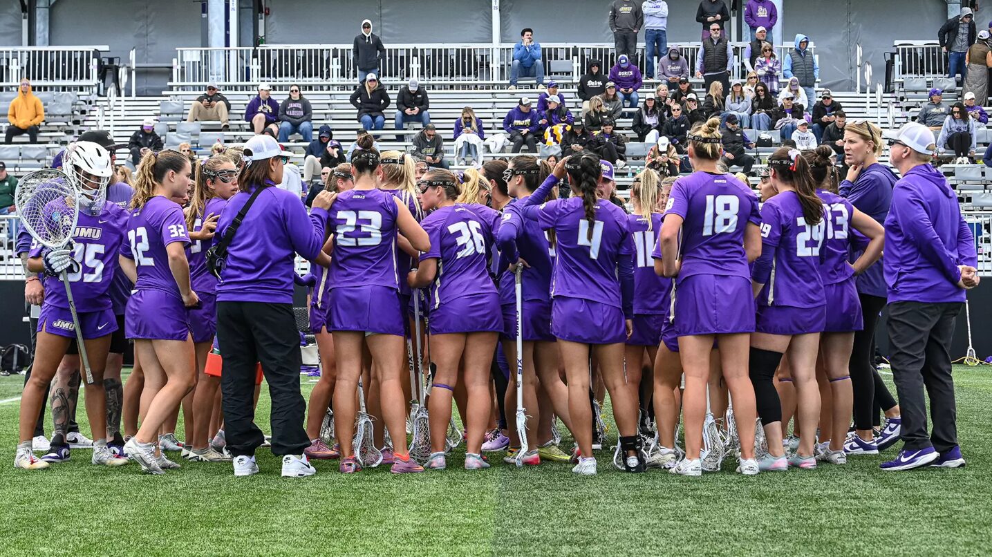 Lacrosse team in purple uniforms huddles on the field with sticks, players leaning in for a pep talk as spectators watch nearby.
