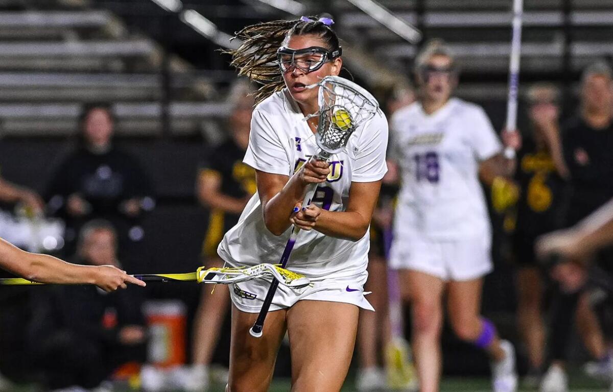 Female lacrosse player in white, goggles on, cradling the ball with her stick while driving forward in a game.