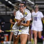 Female lacrosse player in white, goggles on, cradling the ball with her stick while driving forward in a game.