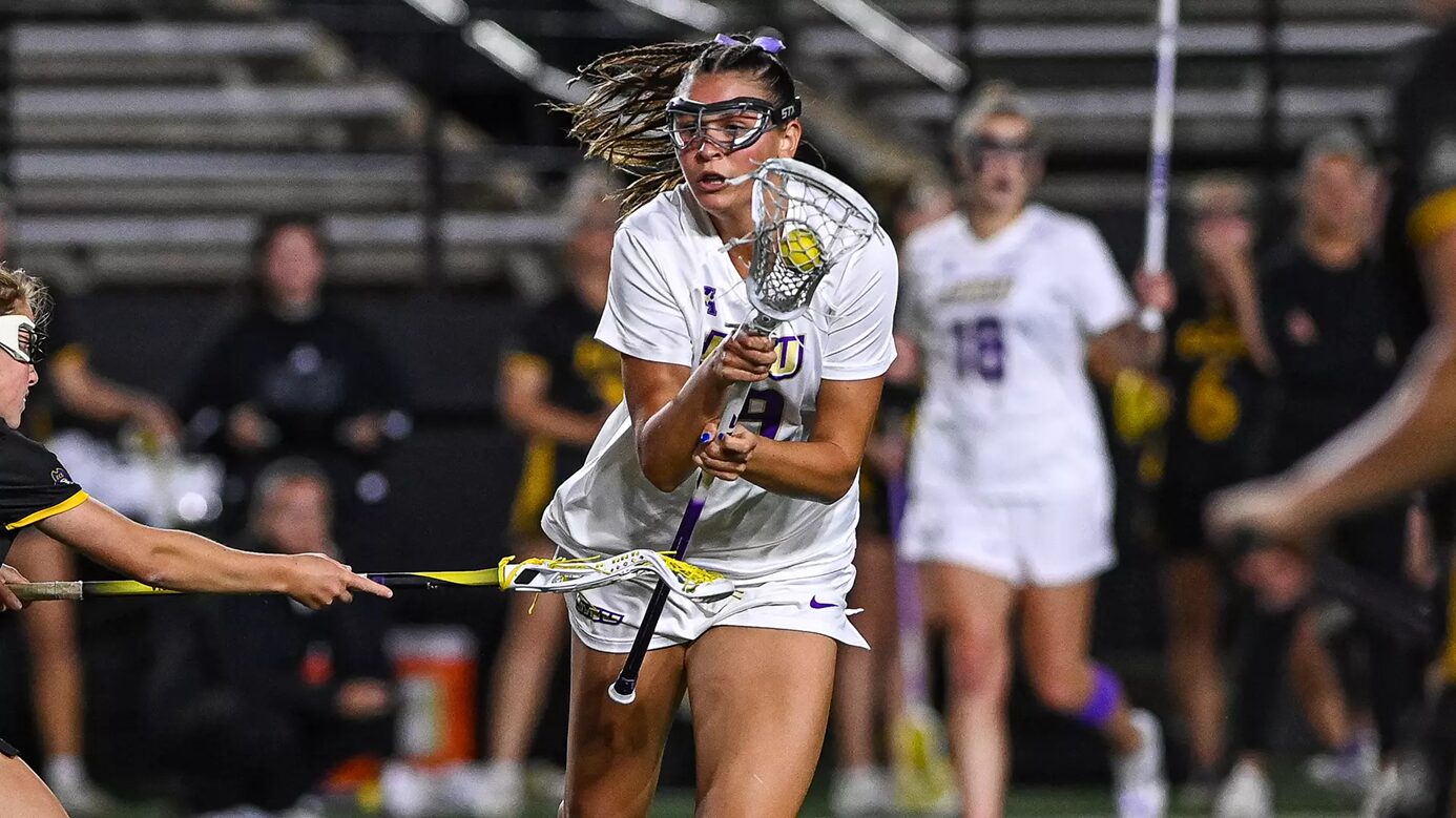 Female lacrosse player in white, goggles on, cradling the ball with her stick while driving forward in a game.