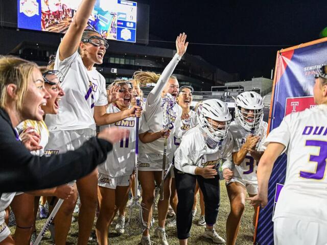 Dukes lacrosse team celebrates a victory on the field at night, players in white uniforms cheering and high-fiving.