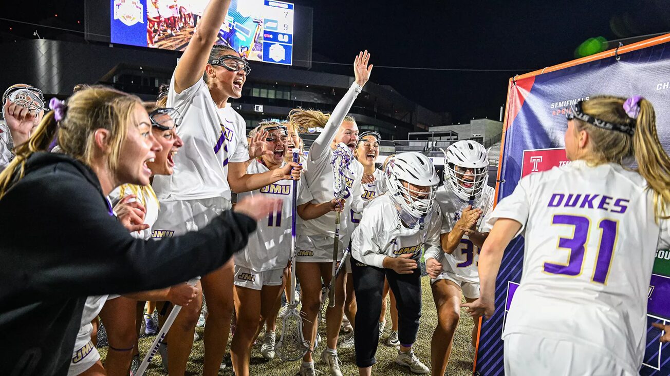 Dukes lacrosse team celebrates a victory on the field at night, players in white uniforms cheering and high-fiving.
