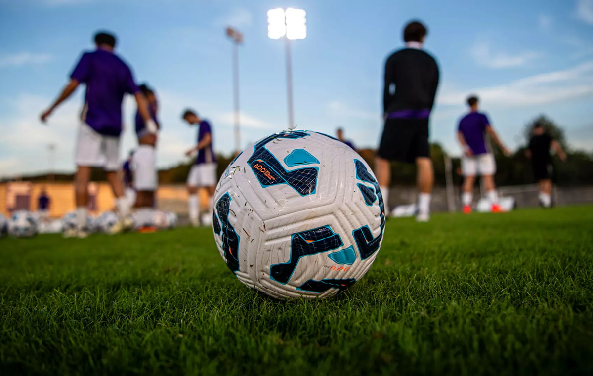 Soccer ball on a green field with players practicing in the background under stadium lights.
