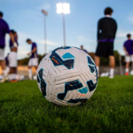 Soccer ball on a green field with players practicing in the background under stadium lights.
