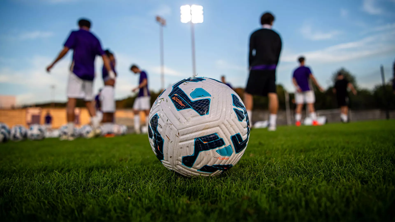 Soccer ball on a green field with players practicing in the background under stadium lights.