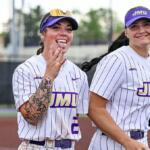 Two JMU softball players in pinstriped uniforms smile at the camera on a field, one showing a hand gesture.