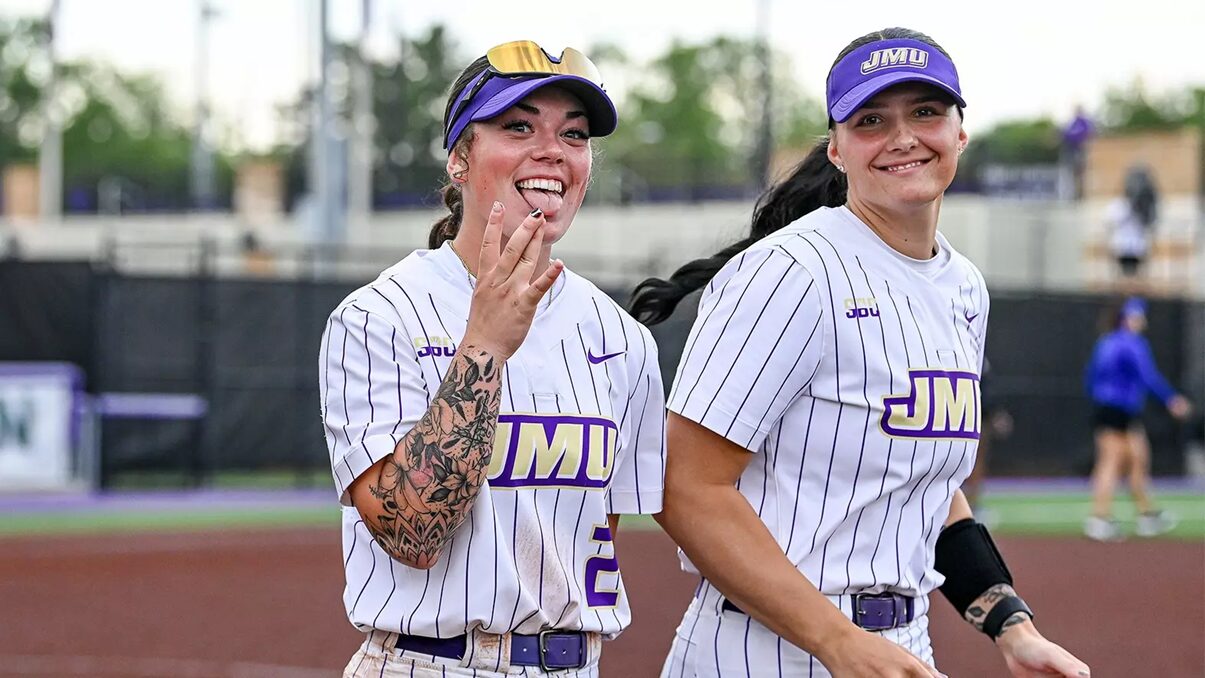 Two JMU softball players in pinstriped uniforms smile at the camera on a field, one showing a hand gesture.