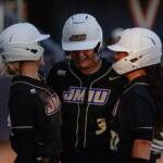 Three JMU softball players in black uniforms and white helmets huddle together on the field.