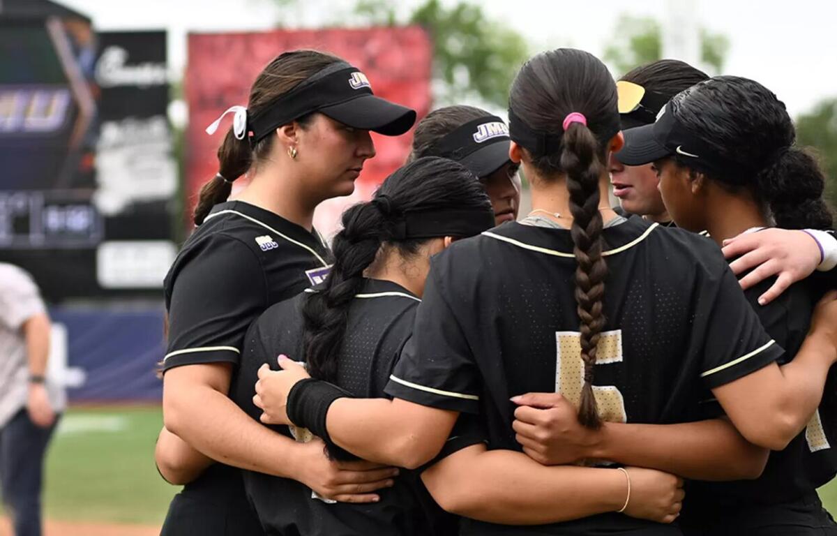 Softball teammates in black uniforms form a huddle with arms around each other on the field.