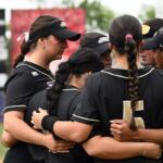 Softball teammates in black uniforms form a huddle with arms around each other on the field.