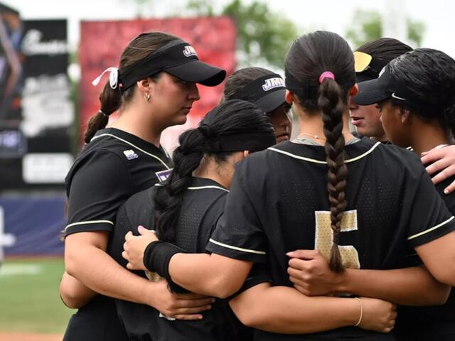 Softball teammates in black uniforms form a huddle with arms around each other on the field.