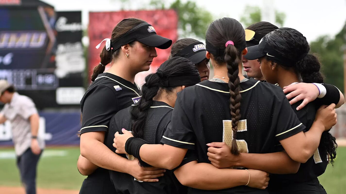 Softball teammates in black uniforms form a huddle with arms around each other on the field.