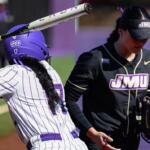 Two softball players collide at the plate: a purple-helmed batter swings while a black-uniform fielder closes in.