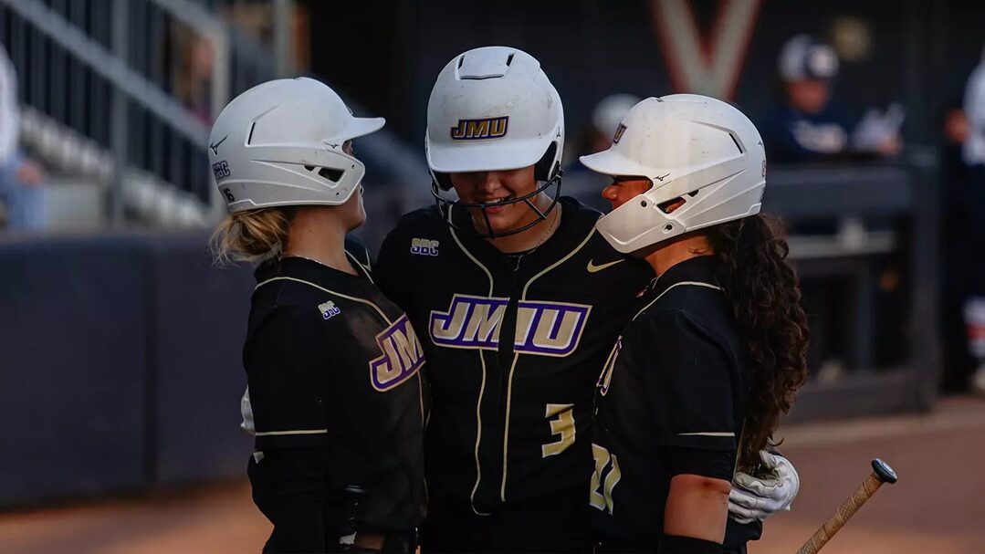 Three JMU softball players in black uniforms and white helmets huddle together on the field.