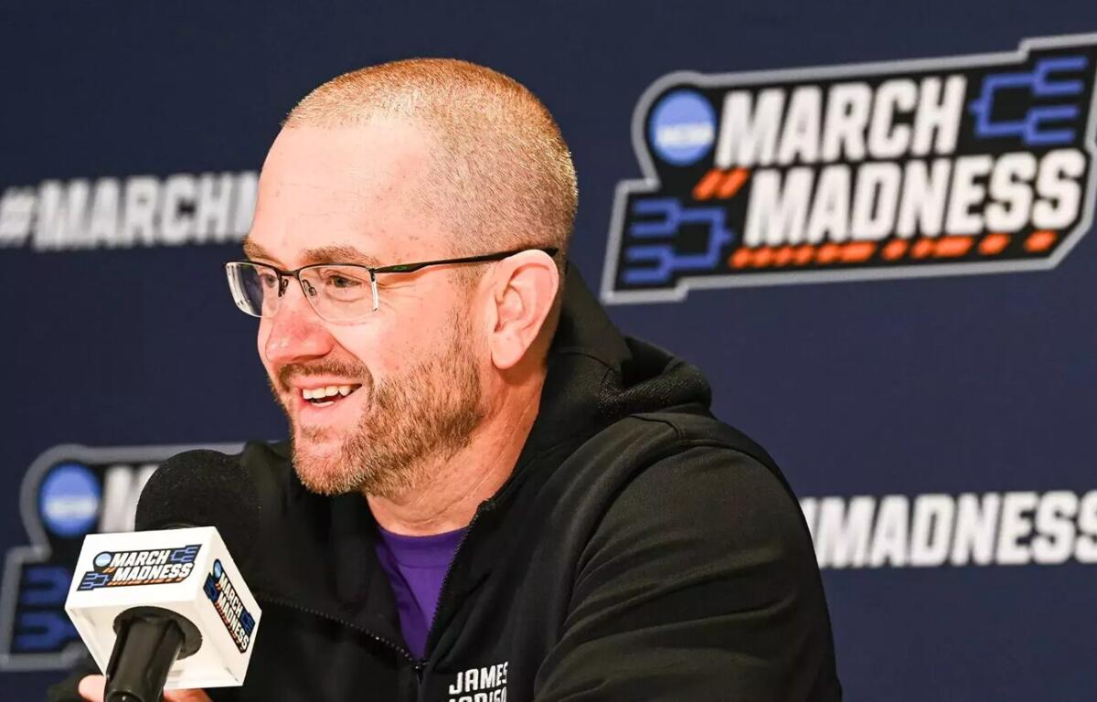Man wearing glasses and a black jacket speaks into a branded microphone at a March Madness press conference, with a blue backdrop and logo behind him.