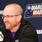 Man wearing glasses and a black jacket speaks into a branded microphone at a March Madness press conference, with a blue backdrop and logo behind him.