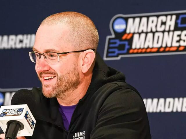Man wearing glasses and a black jacket speaks into a branded microphone at a March Madness press conference, with a blue backdrop and logo behind him.