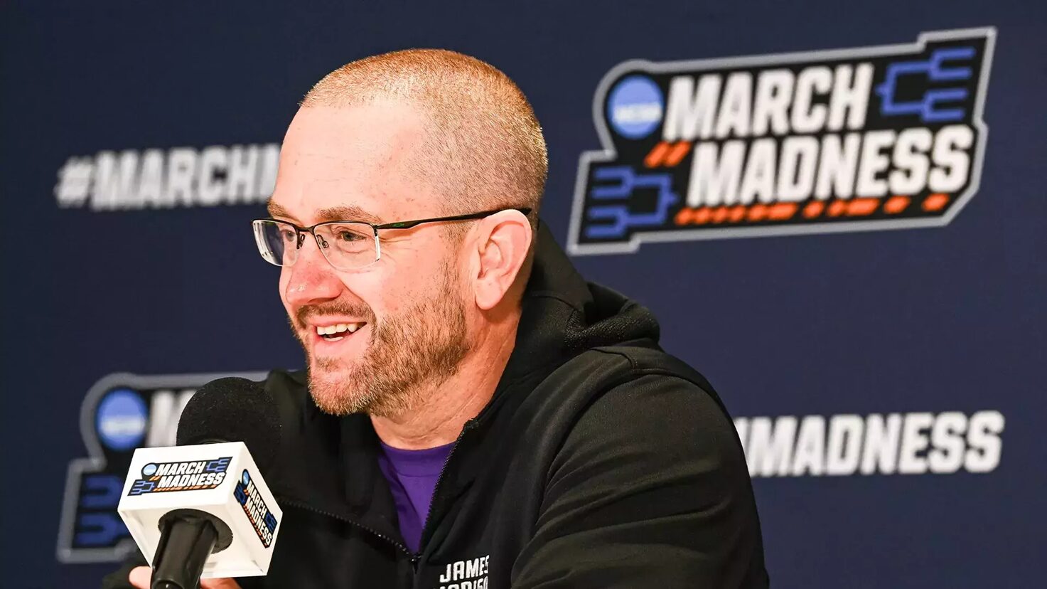 Man wearing glasses and a black jacket speaks into a branded microphone at a March Madness press conference, with a blue backdrop and logo behind him.