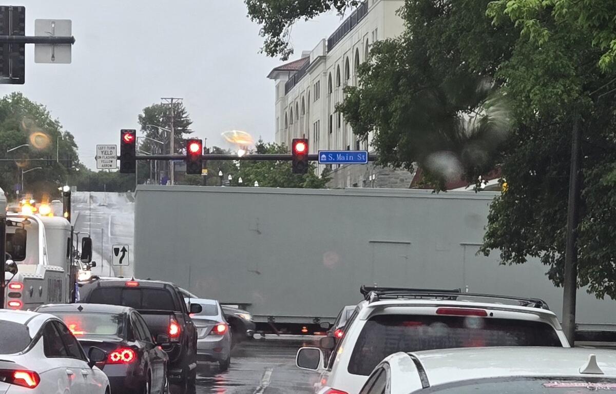 Street in heavy traffic on a rainy day, a long gray trailer blocks part of the lane near a blue S. Main St sign and red traffic lights overhead.