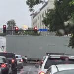 Street in heavy traffic on a rainy day, a long gray trailer blocks part of the lane near a blue S. Main St sign and red traffic lights overhead.