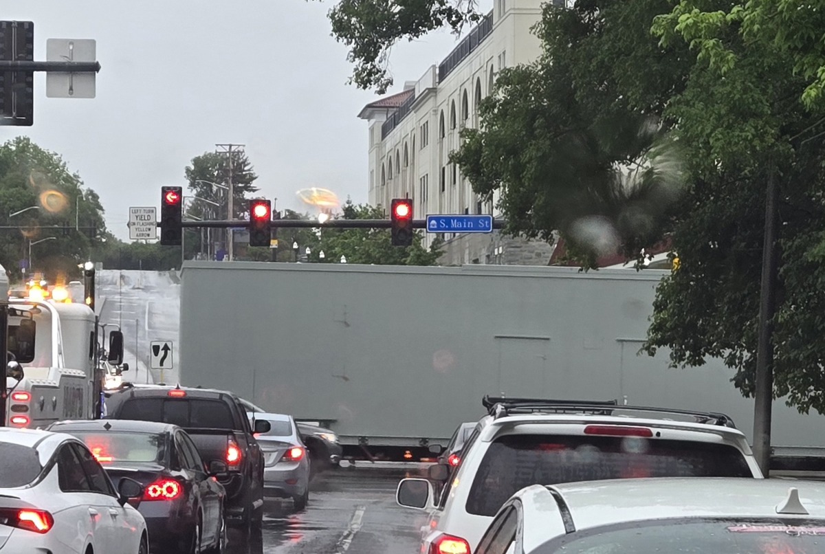 Street in heavy traffic on a rainy day, a long gray trailer blocks part of the lane near a blue S. Main St sign and red traffic lights overhead.