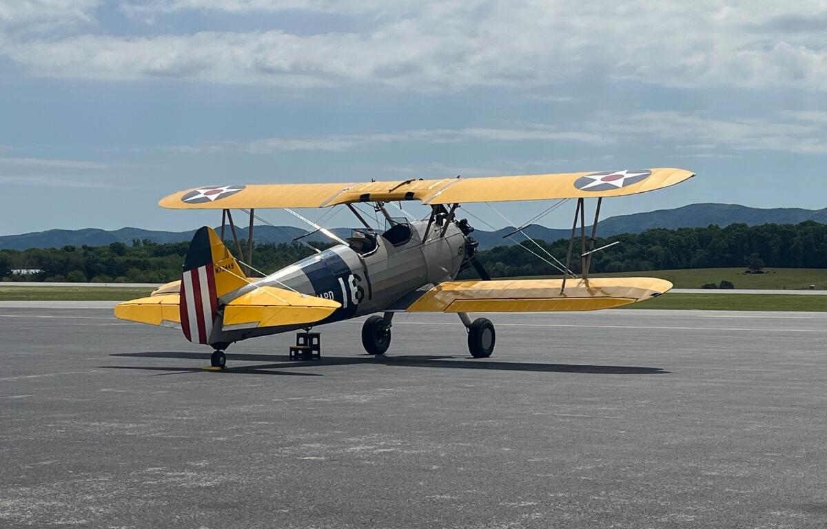 Yellow World War I–era biplane taxiing on a runway, silver fuselage with U.S. roundel insignia and a striped red‑white tail.