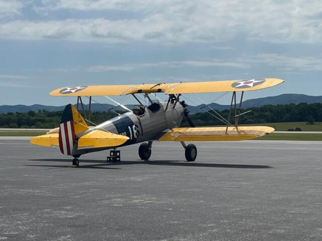 Yellow World War I–era biplane taxiing on a runway, silver fuselage with U.S. roundel insignia and a striped red‑white tail.