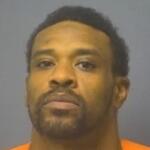 Mugshot portrait of a man with short dark hair and facial hair wearing an orange jail jumpsuit against a gray backdrop, looking at the camera.