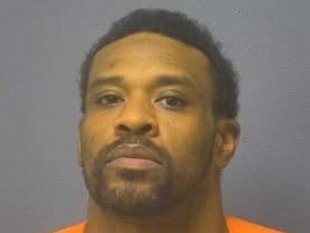 Mugshot portrait of a man with short dark hair and facial hair wearing an orange jail jumpsuit against a gray backdrop, looking at the camera.