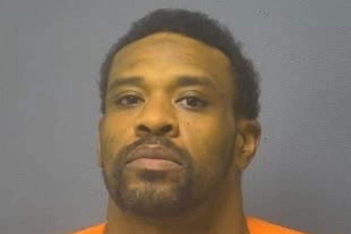 Mugshot portrait of a man with short dark hair and facial hair wearing an orange jail jumpsuit against a gray backdrop, looking at the camera.