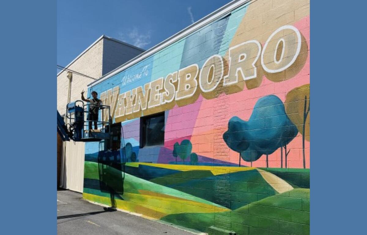 Worker on a lift painting a colorful mural on a brick wall that reads 'WAYNESBORO'.