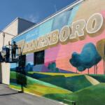 Worker on a lift painting a colorful mural on a brick wall that reads 'WAYNESBORO'.